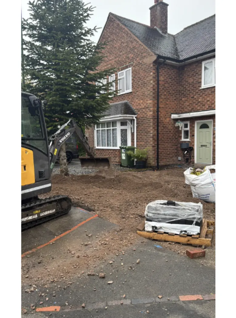 Mini digger excavating a front garden for a new driveway, pallet of blocks and a builders bag ready beside a brick semi